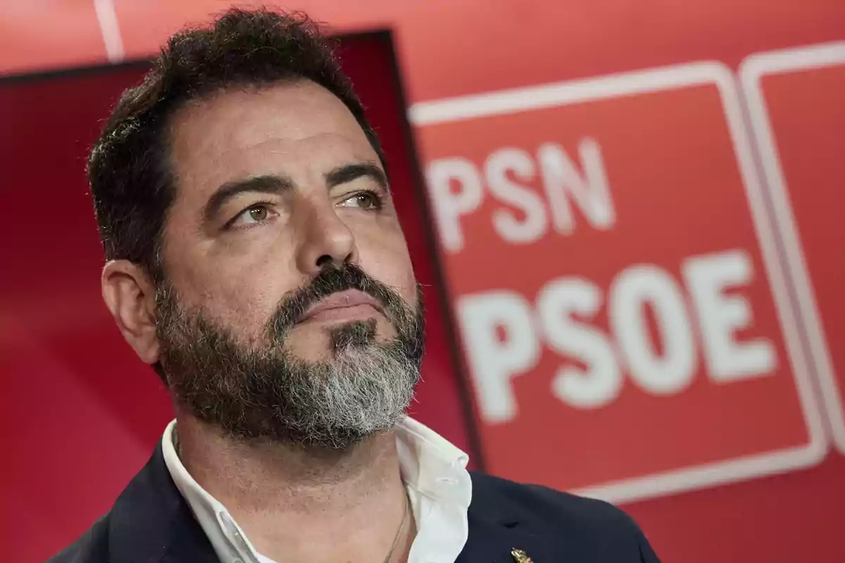 Hombre de barba y cabello oscuro con expresión seria frente a un fondo rojo con logotipos de PSN y PSOE desenfocados