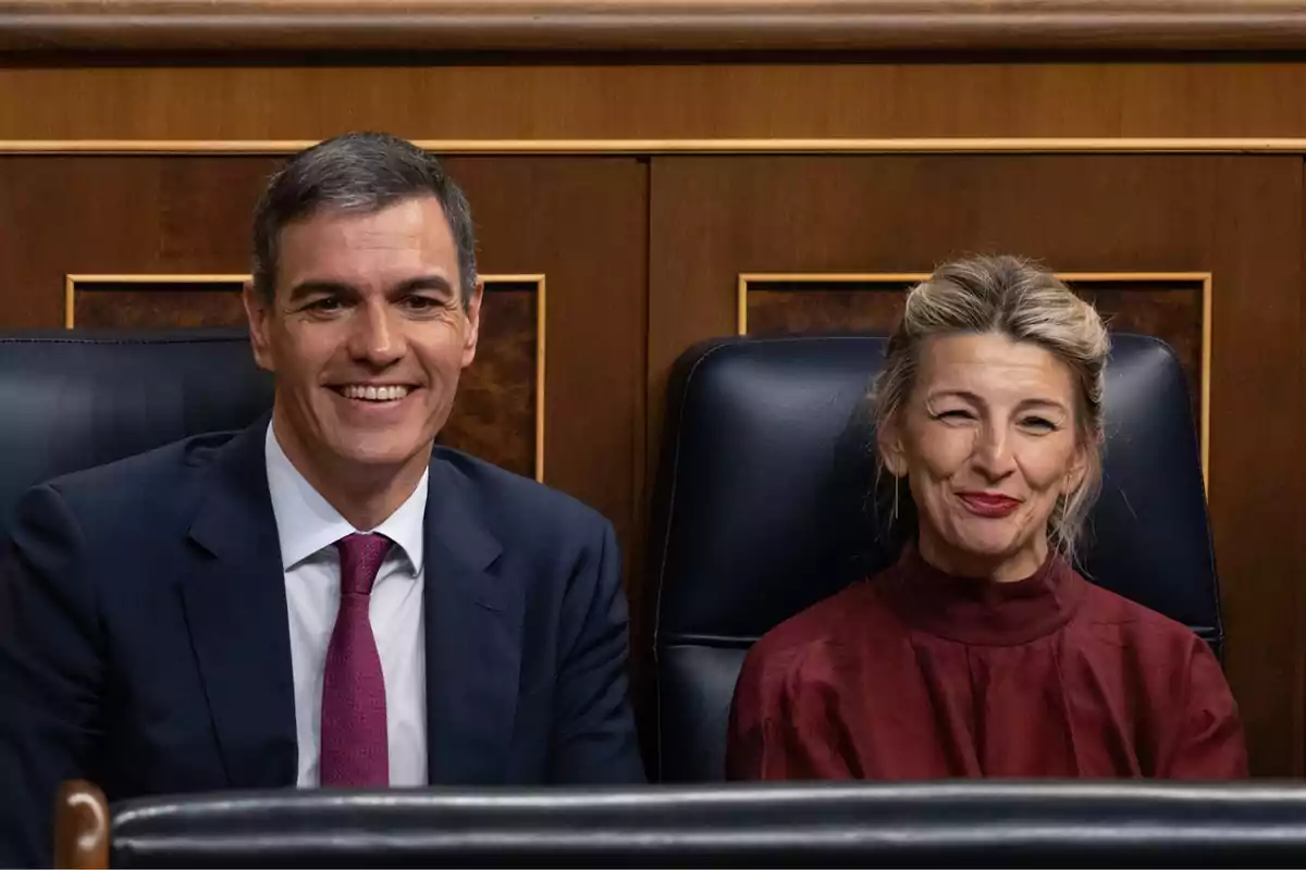 Dos personas sentadas en sillas de cuero negro en un entorno formal, una de ellas con traje y corbata roja y la otra con blusa roja.