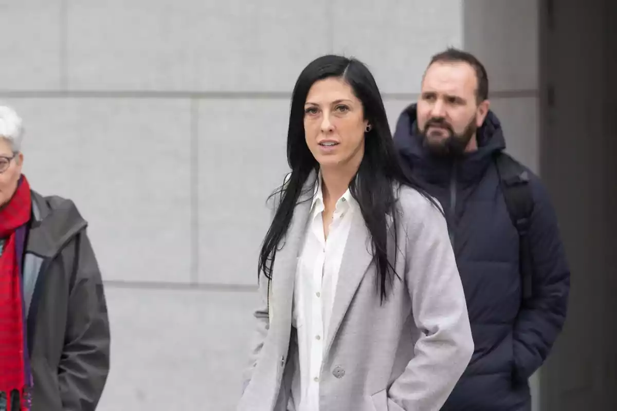 A woman with dark hair and a gray coat walks outdoors accompanied by two people.