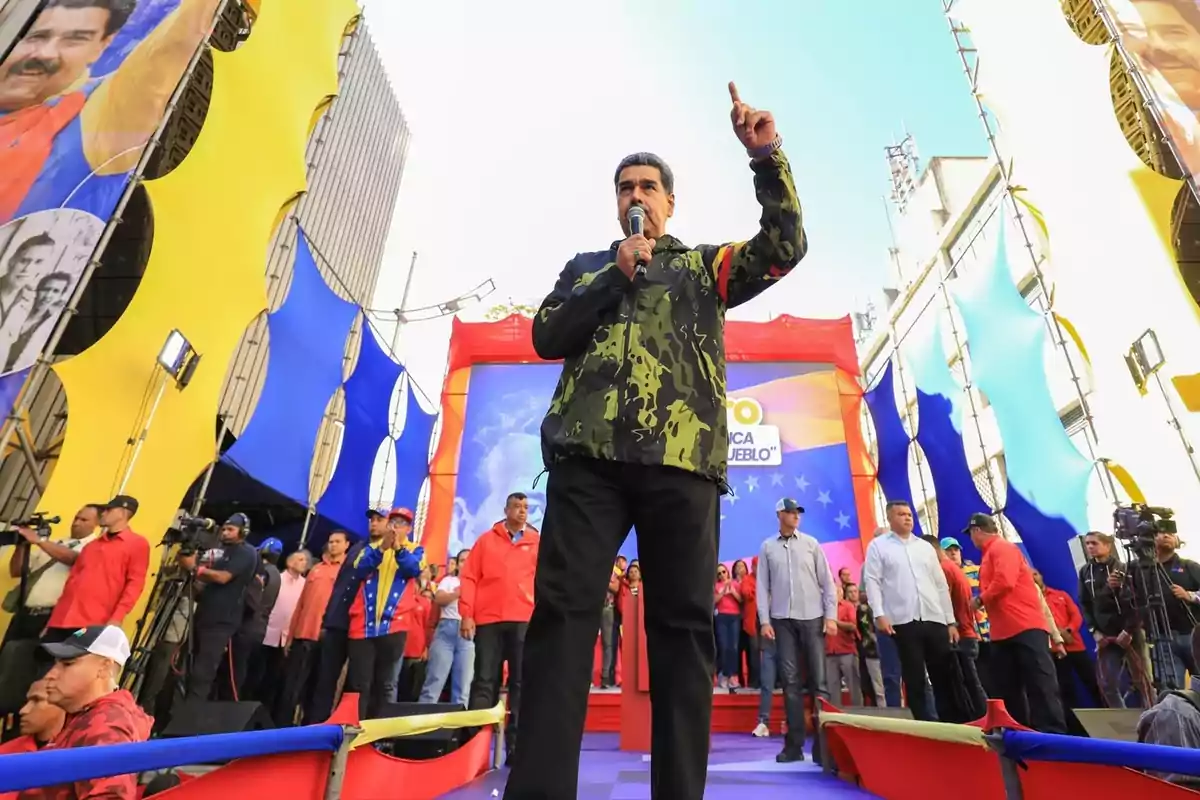 Un hombre con chaqueta de camuflaje habla en un mitin político, rodeado de personas y banderas de colores.