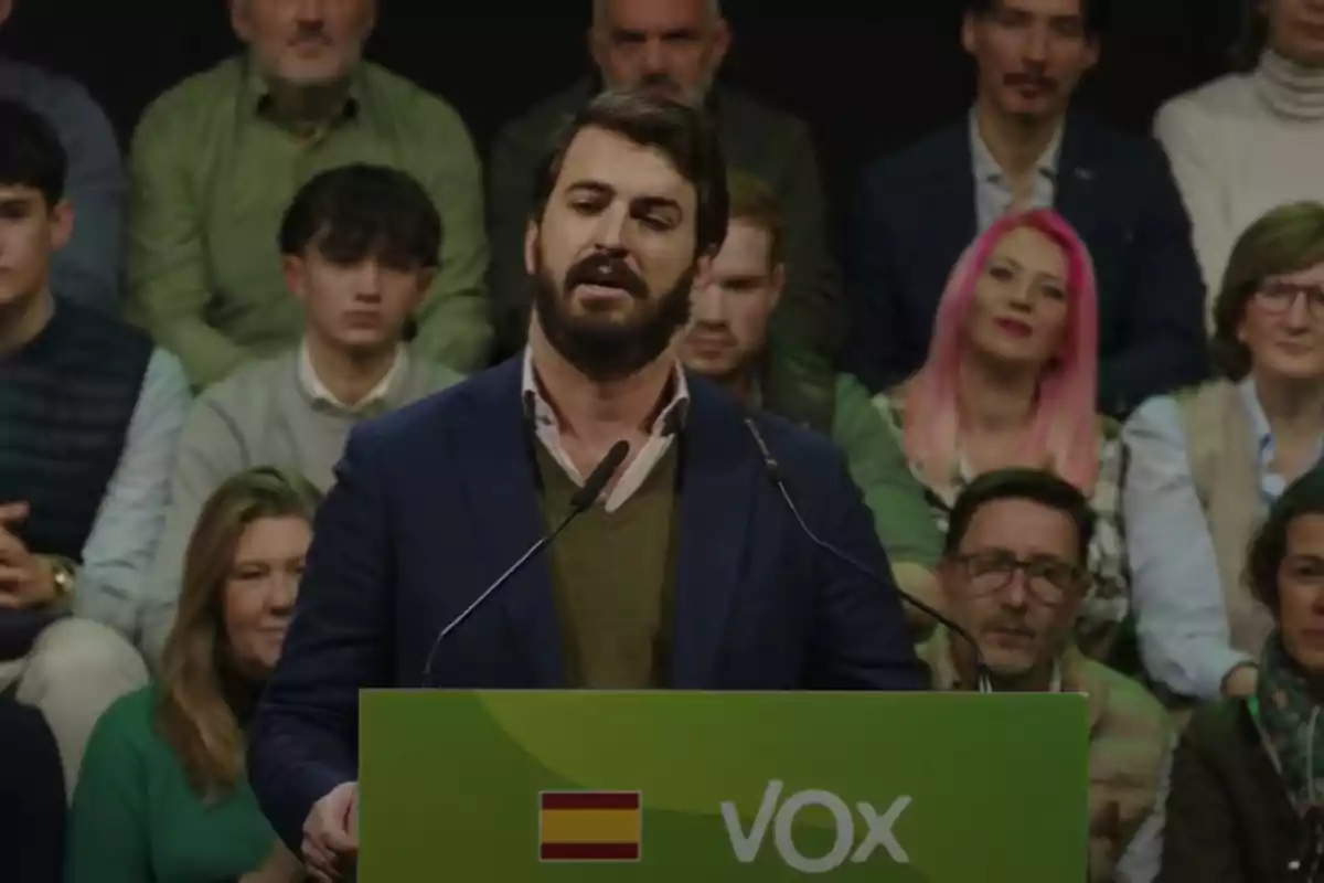 A man speaking at a podium with a microphone, surrounded by seated people, with a green sign featuring a flag and the text "VOX".