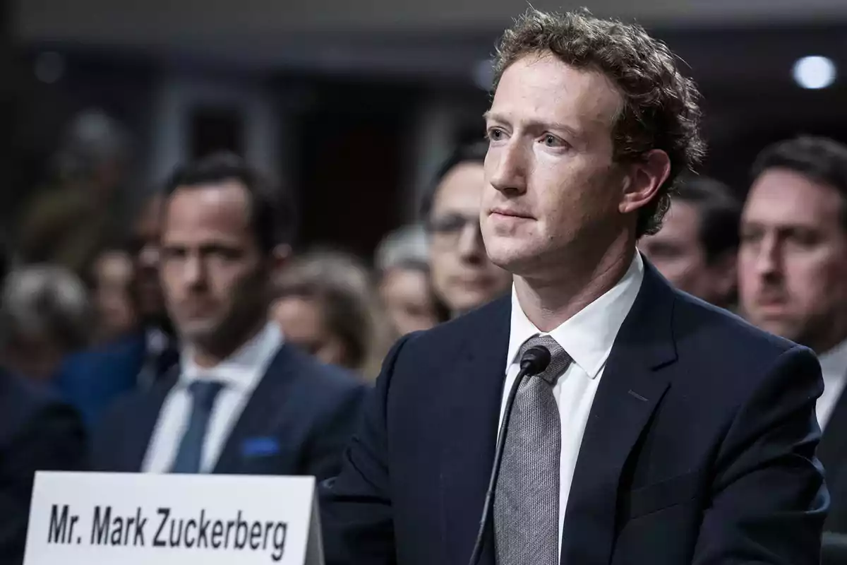 Un hombre en traje oscuro y corbata gris, sentado frente a un micrófono con un cartel que dice "Mr. Mark Zuckerberg", rodeado de personas en un entorno formal.