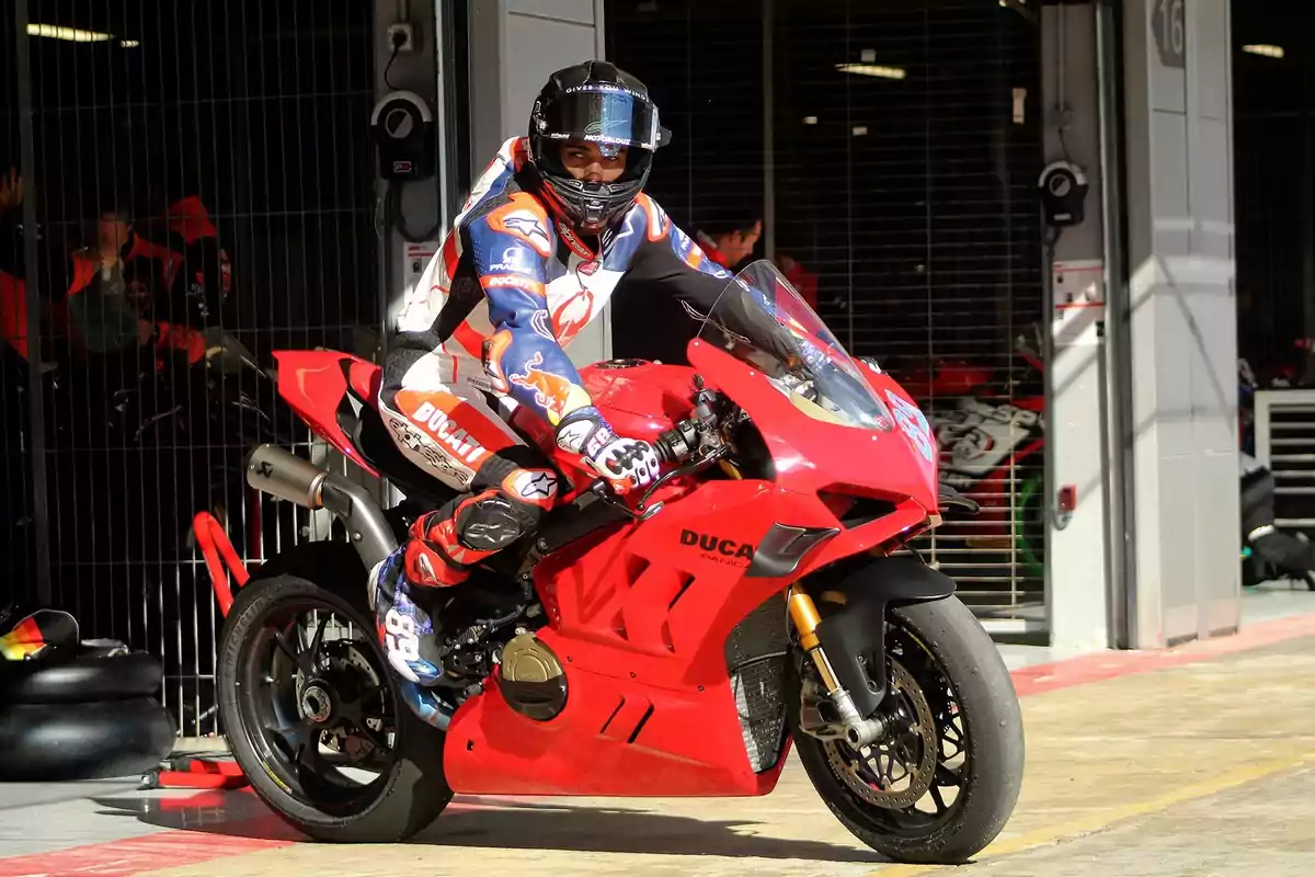 Piloto de motociclismo con traje de carreras y casco, montando una motocicleta Ducati roja en un garaje de boxes.