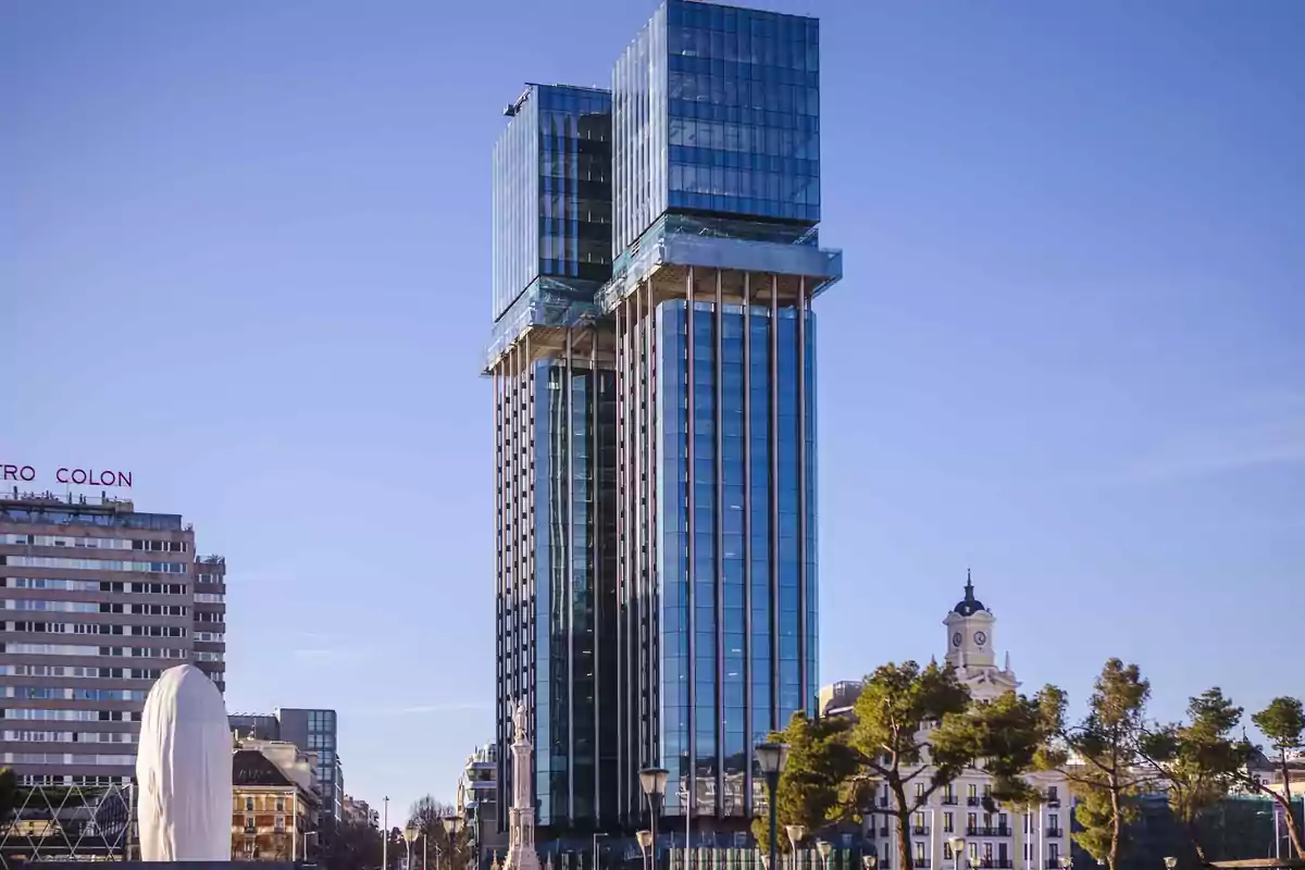 Edificio alto y moderno con fachada de vidrio en una ciudad, rodeado de otros edificios y árboles.