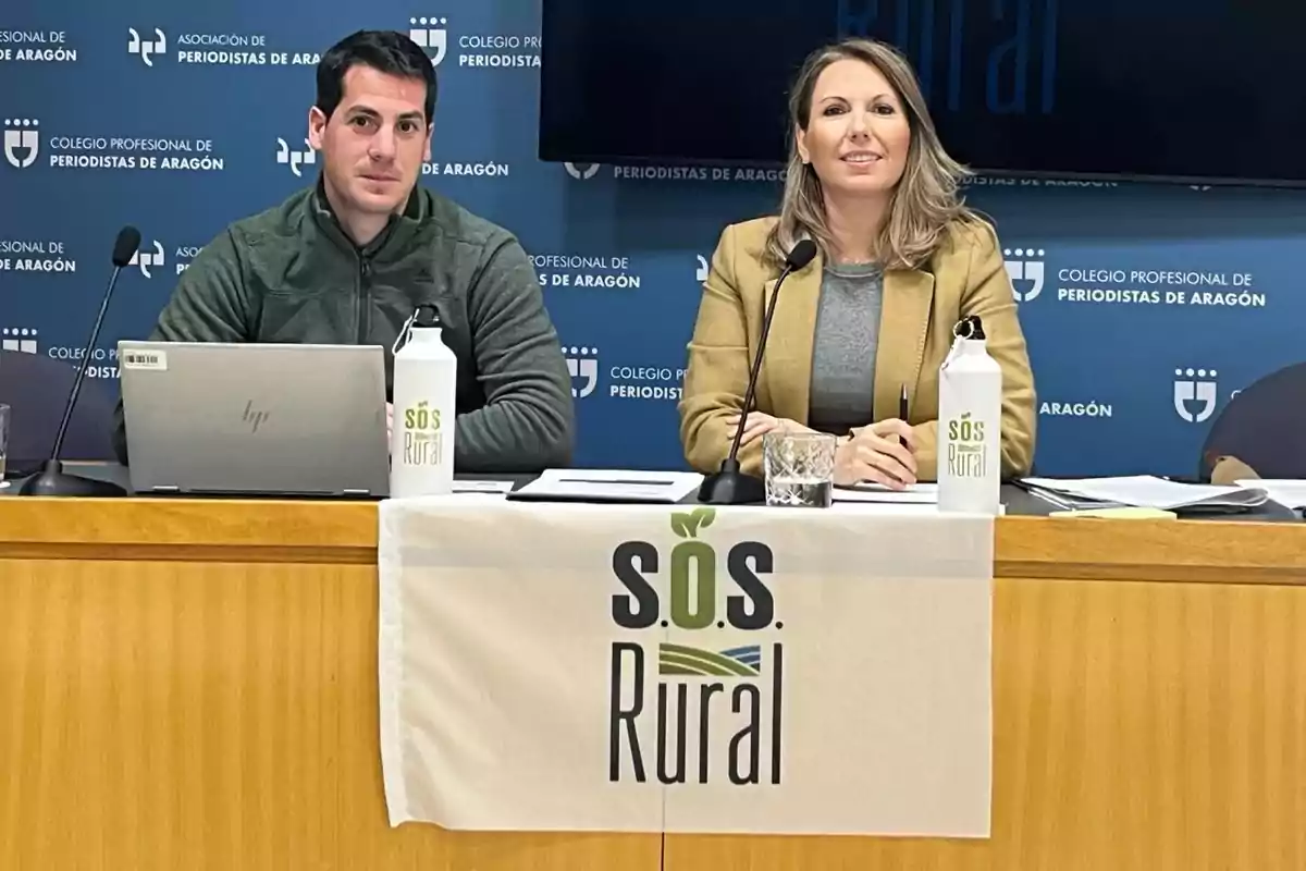 Dos personas sentadas en una mesa con un cartel que dice "S.O.S. Rural" en un evento del Colegio Profesional de Periodistas de Aragón.