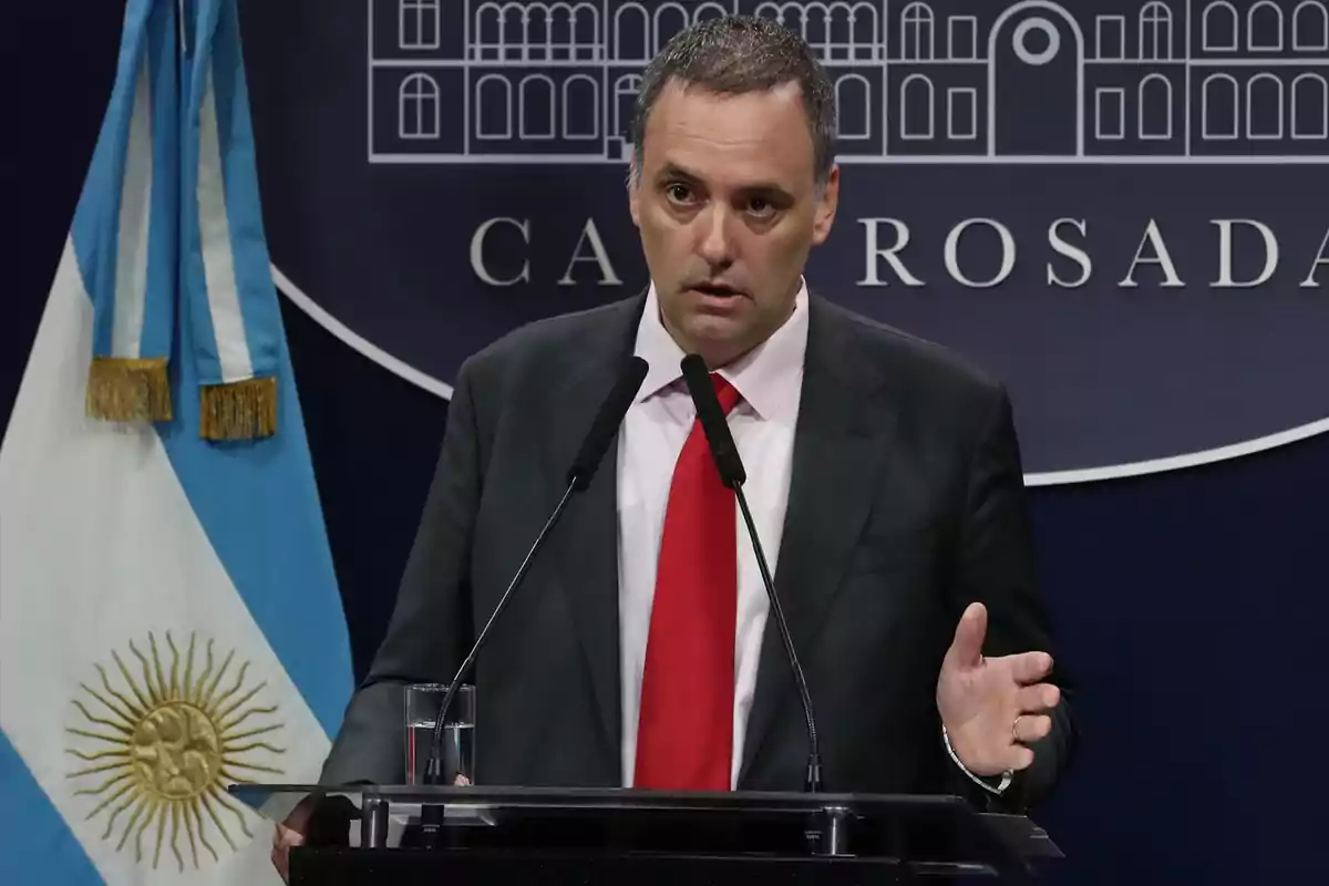 Un hombre de traje y corbata roja hablando en una conferencia de prensa con una bandera argentina y un cartel de la Casa Rosada detrás.