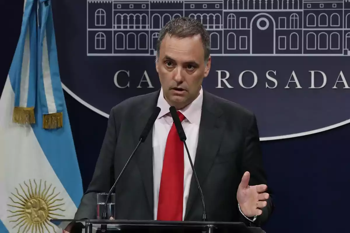 Un hombre de traje y corbata roja habla en un podio con dos micrófonos, con una bandera argentina y un fondo que dice "Casa Rosada".