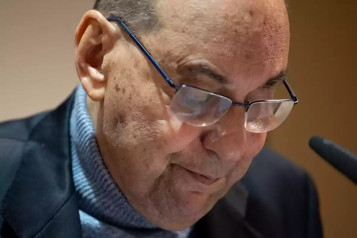 Un hombre mayor con gafas y suéter gris, mirando hacia abajo con expresión serena.