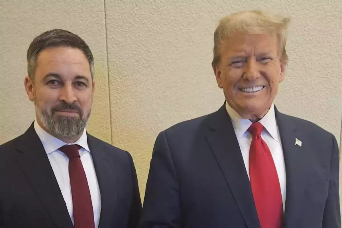 Two men in dark suits and red ties pose together smiling in front of a light-colored wall.