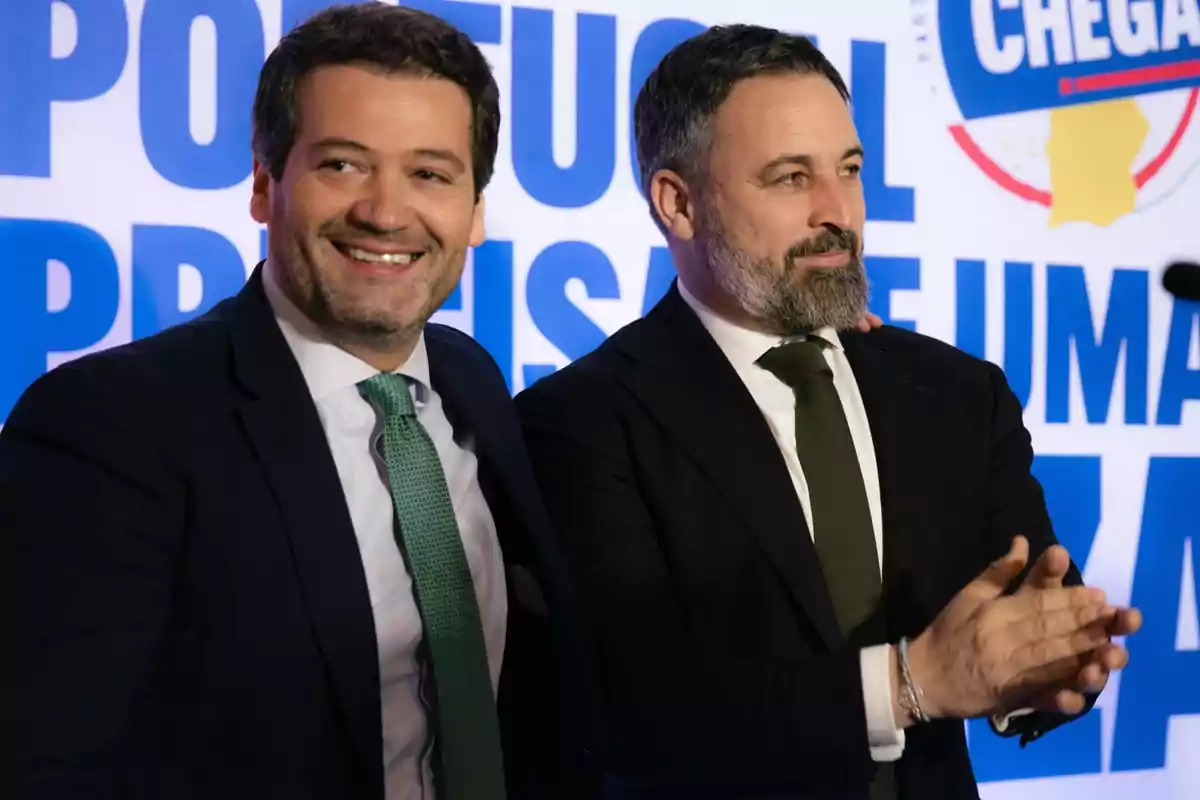 Dos hombres con trajes oscuros y corbatas verdes sonríen frente a un fondo con texto azul y un logotipo circular.