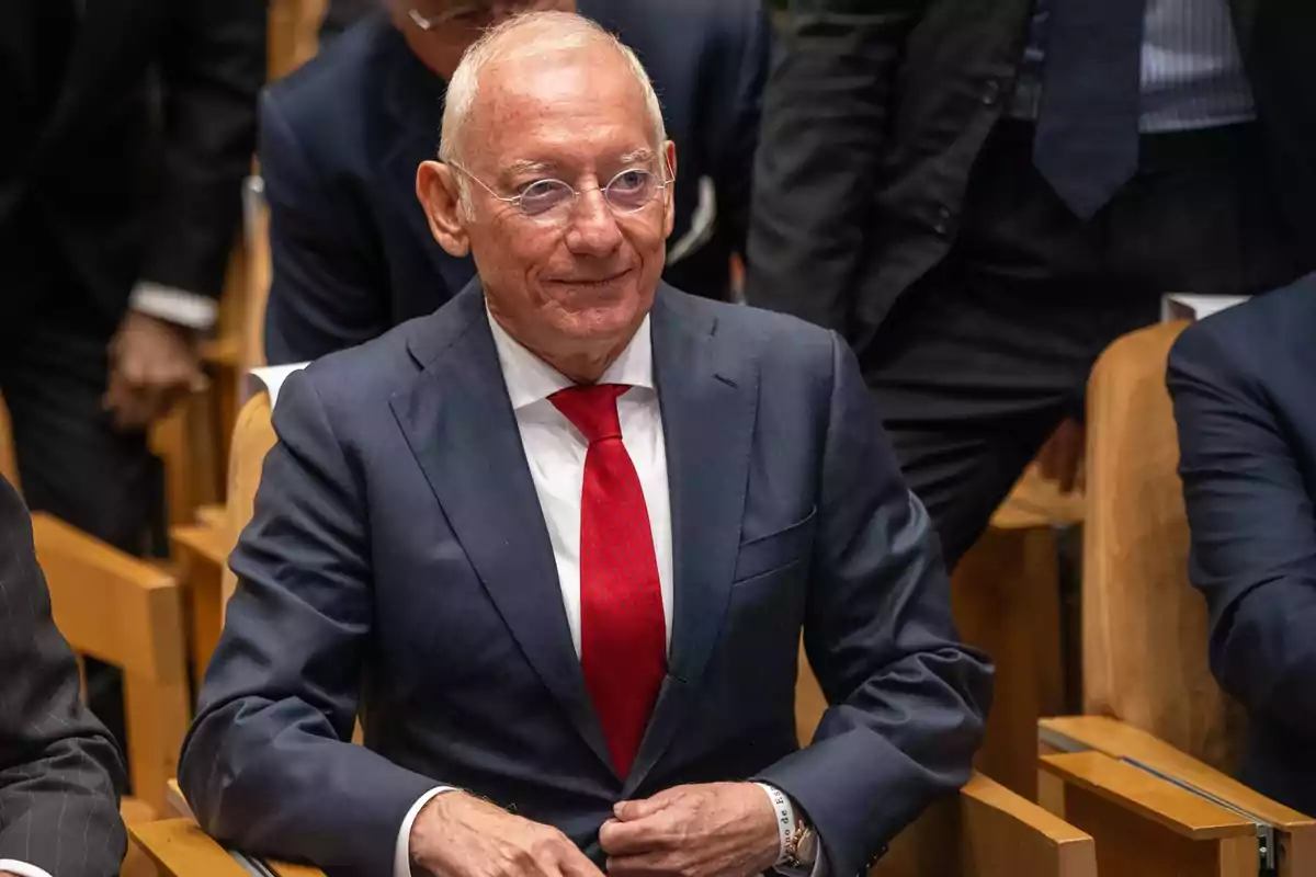 Un hombre mayor con gafas y traje oscuro con corbata roja sentado en una sala de conferencias.