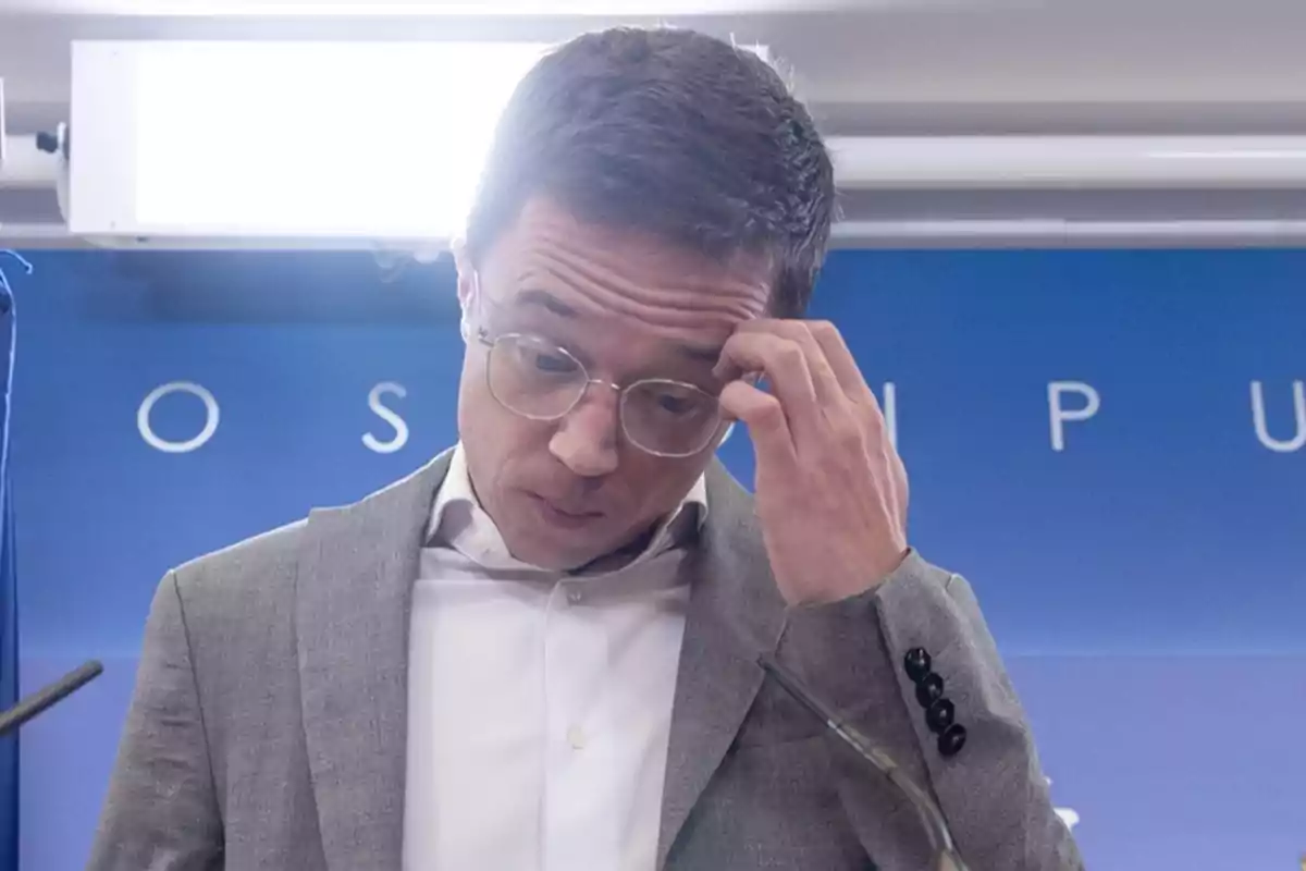 Un hombre con gafas y traje gris se toca la cabeza mientras mira hacia abajo en un entorno de conferencia.