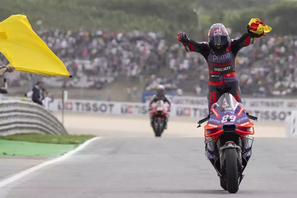 Un motociclista celebra mientras realiza un caballito en su moto de carreras, sosteniendo una bandera en la mano, con una multitud de espectadores al fondo.