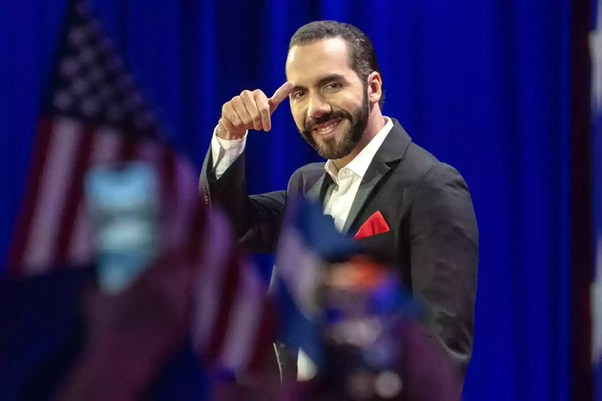 Hombre con traje oscuro y pañuelo rojo en el bolsillo, sonriendo y señalando con el dedo índice hacia su cabeza, con banderas desenfocadas en primer plano y un fondo azul.