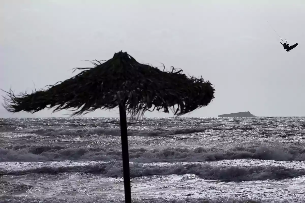 Una sombrilla de paja en la playa con el mar agitado y una persona practicando kitesurf en el fondo.