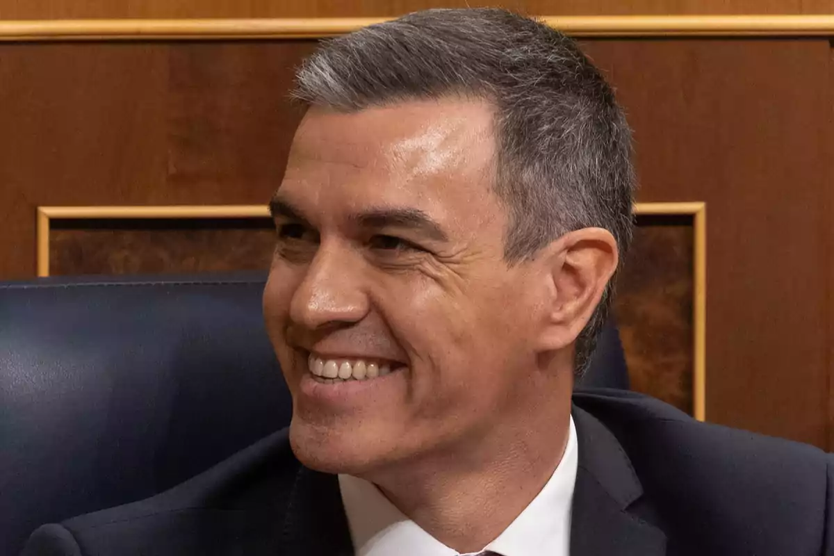 Un hombre de cabello canoso y traje oscuro sonríe mientras está sentado en un entorno formal. Un hombre de cabello canoso y traje oscuro sonríe mientras está sentado en un entorno formal.