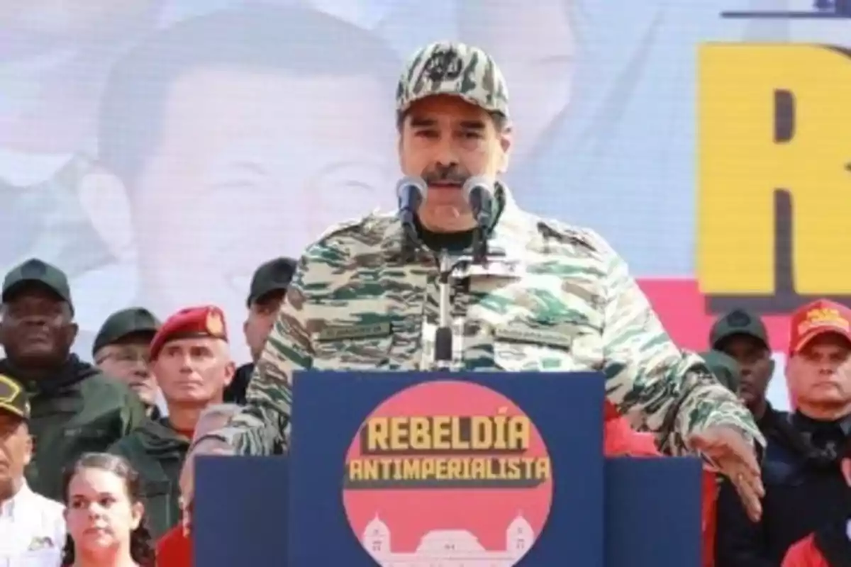 Un hombre con uniforme militar habla en un podio con un cartel que dice "Rebeldía Antiimperialista", rodeado de personas también en uniforme.