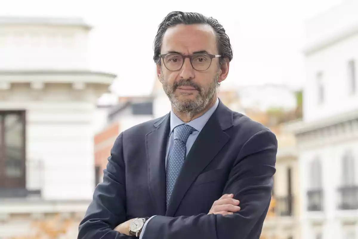 Hombre con traje y gafas de pie con los brazos cruzados en un entorno urbano.