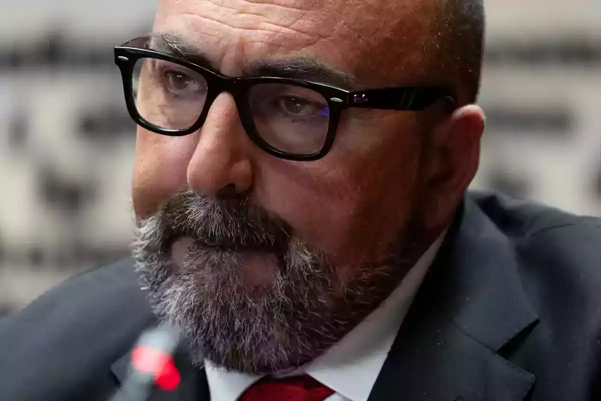 Un hombre con barba y gafas negras, vestido con traje oscuro y corbata roja, mirando pensativamente.