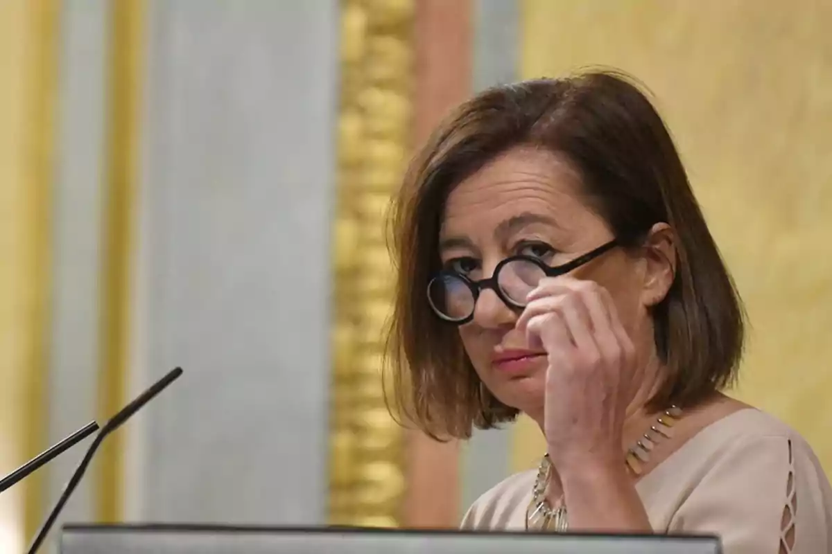 Una mujer con gafas redondas ajustándose las lentes mientras está de pie frente a un micrófono en un entorno formal.