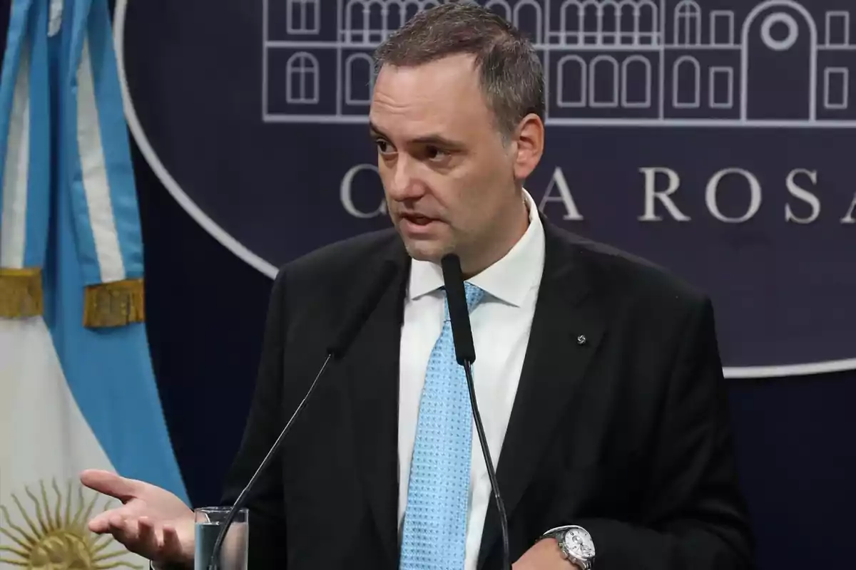 Hombre de traje y corbata azul hablando en una conferencia de prensa con la bandera de Argentina en el fondo.