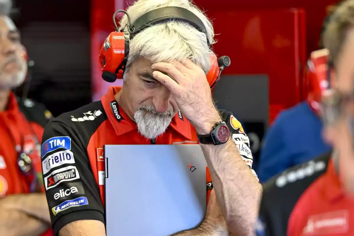 Un hombre con auriculares rojos y uniforme de equipo de carreras sostiene una carpeta mientras se toca la frente con una expresión de preocupación.