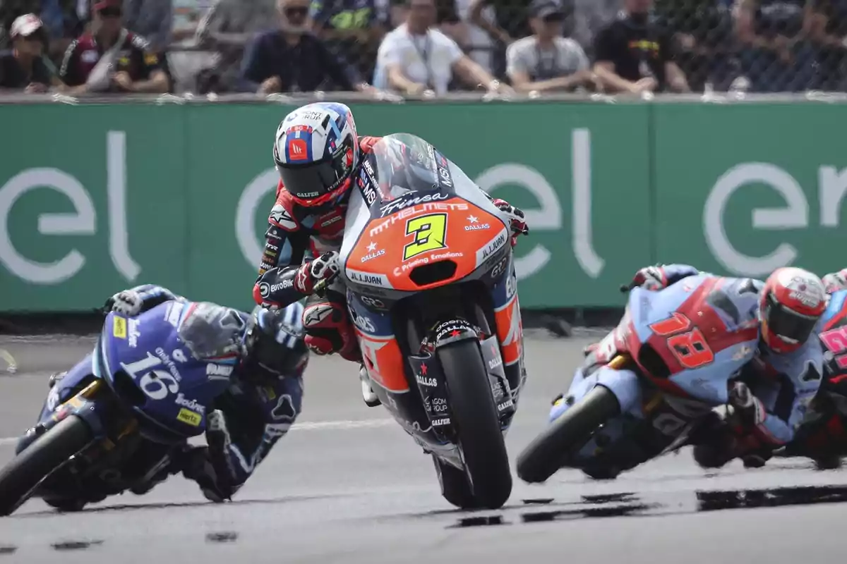 Tres motociclistas compiten en una carrera de motos, con espectadores de fondo.