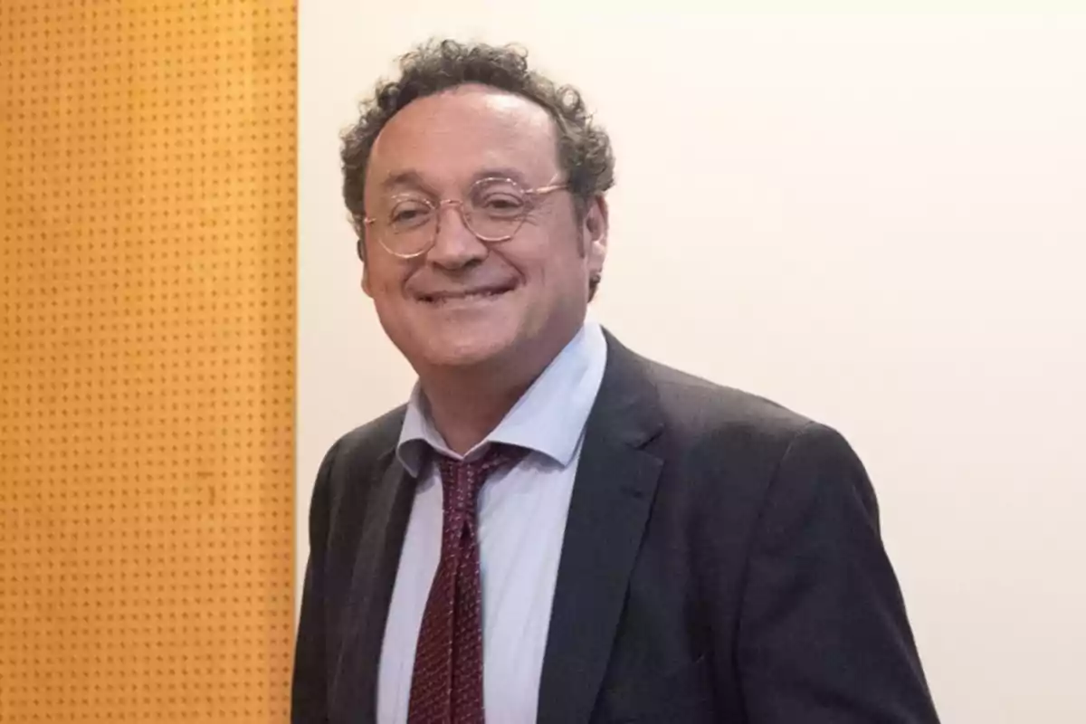 Man in glasses and suit smiles in front of a light wall and a wood paneled wall.