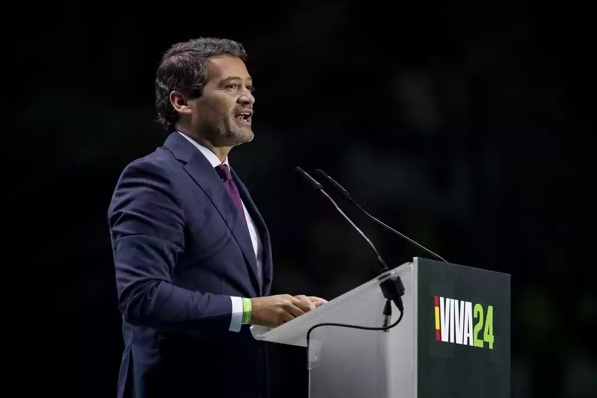 A man in a dark blue suit speaks at a podium with the 