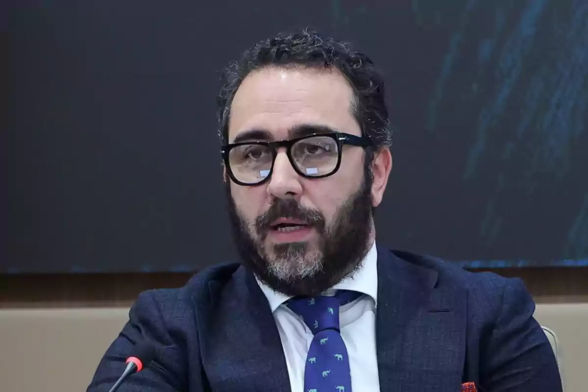 A bearded man with glasses speaks into a microphone during a formal event. A bearded man with glasses speaks into a microphone during a formal event.