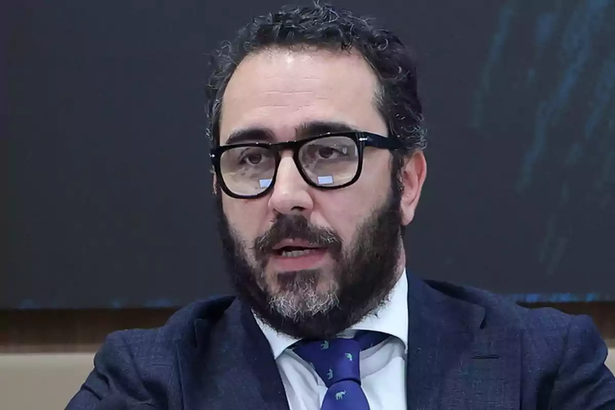 Un hombre con barba y gafas, vistiendo un traje oscuro y corbata azul, hablando frente a un fondo oscuro.