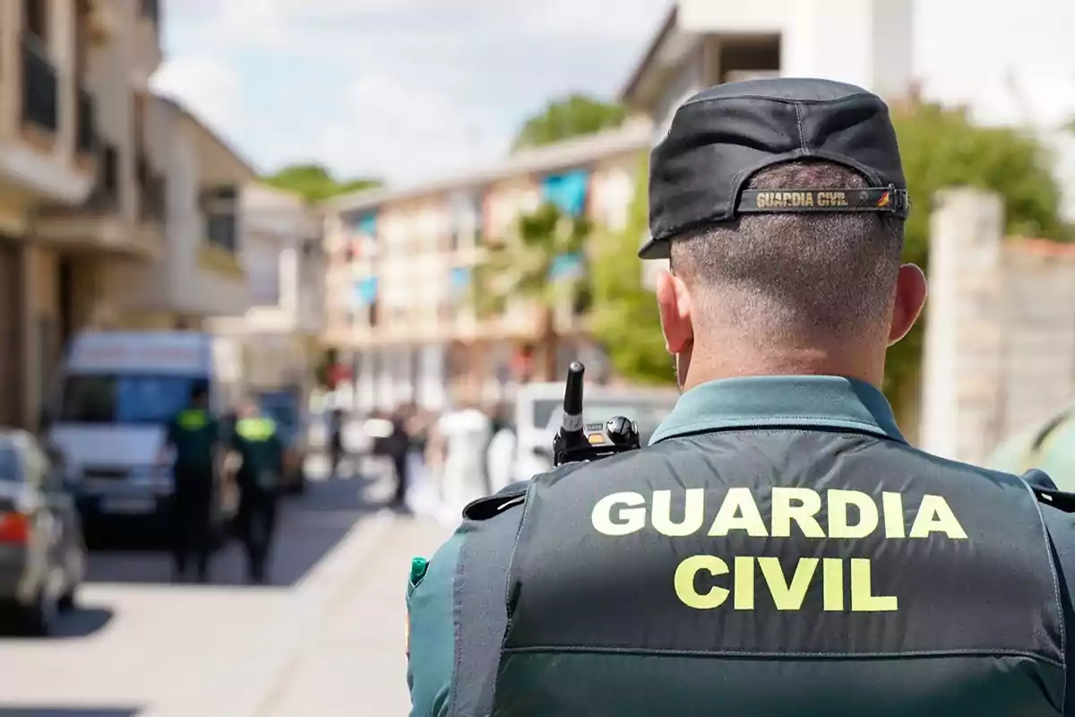 Un agente de la Guardia Civil de espaldas en una calle con otros agentes y vehículos al fondo.