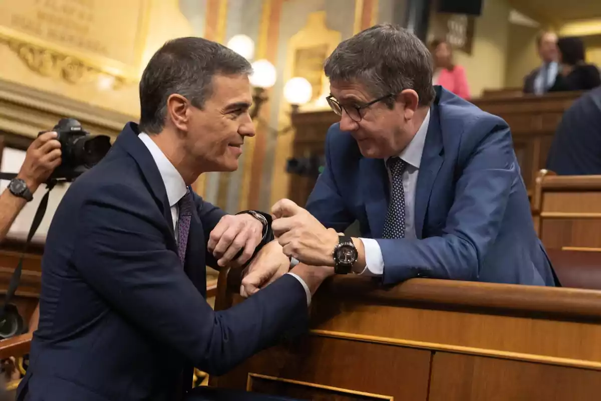 Dos hombres de traje conversan y sonríen en un entorno formal mientras un fotógrafo captura el momento.