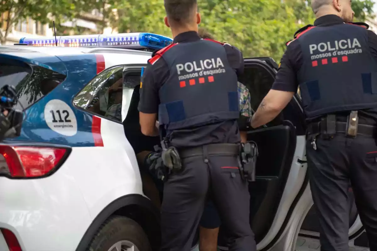 Dos agentes de la policía de los Mossos d'Esquadra están escoltando a una persona hacia el interior de un coche patrulla con las luces encendidas.