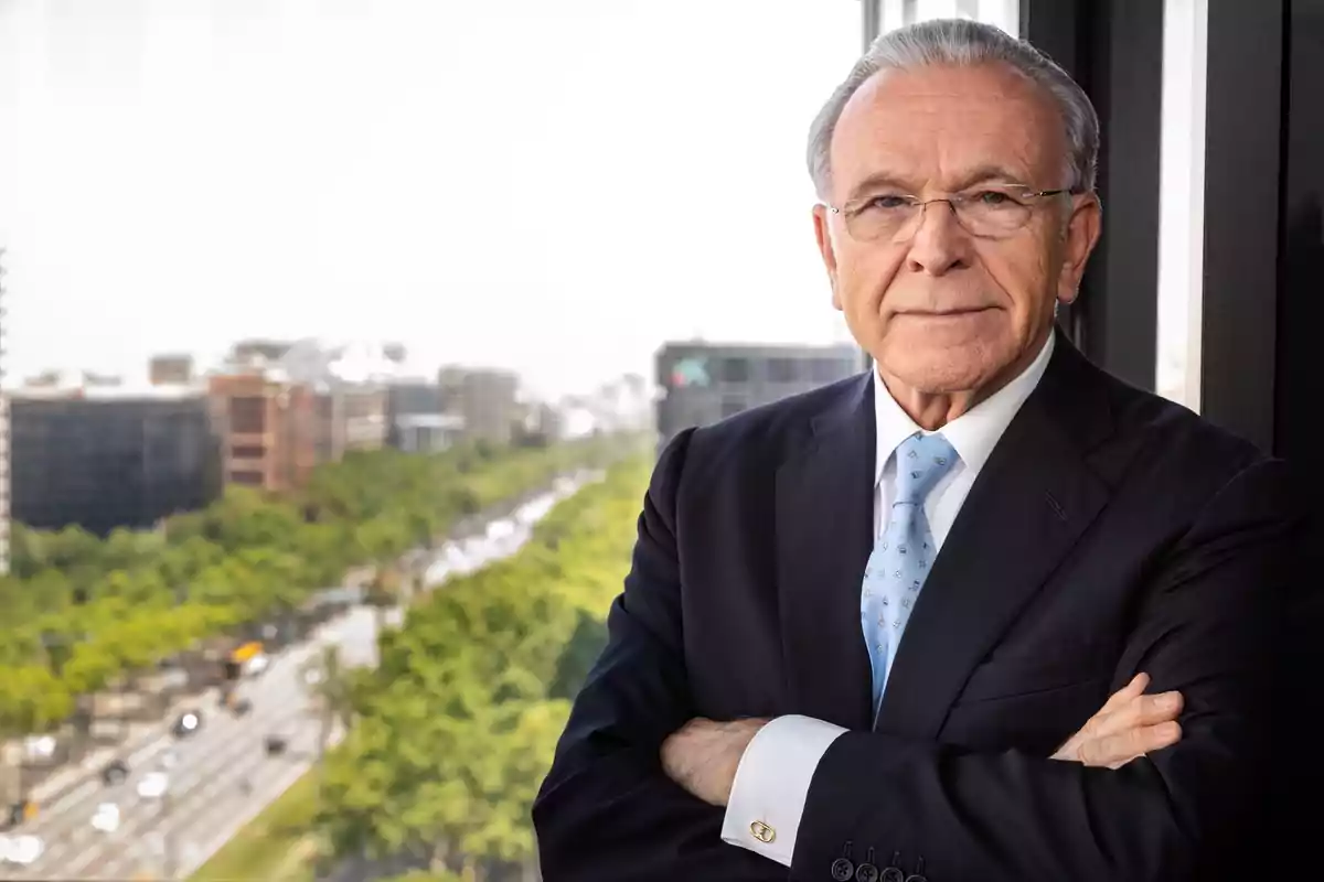 Un hombre mayor con traje y corbata azul claro está de pie con los brazos cruzados frente a una ventana con vista a una avenida arbolada y edificios.