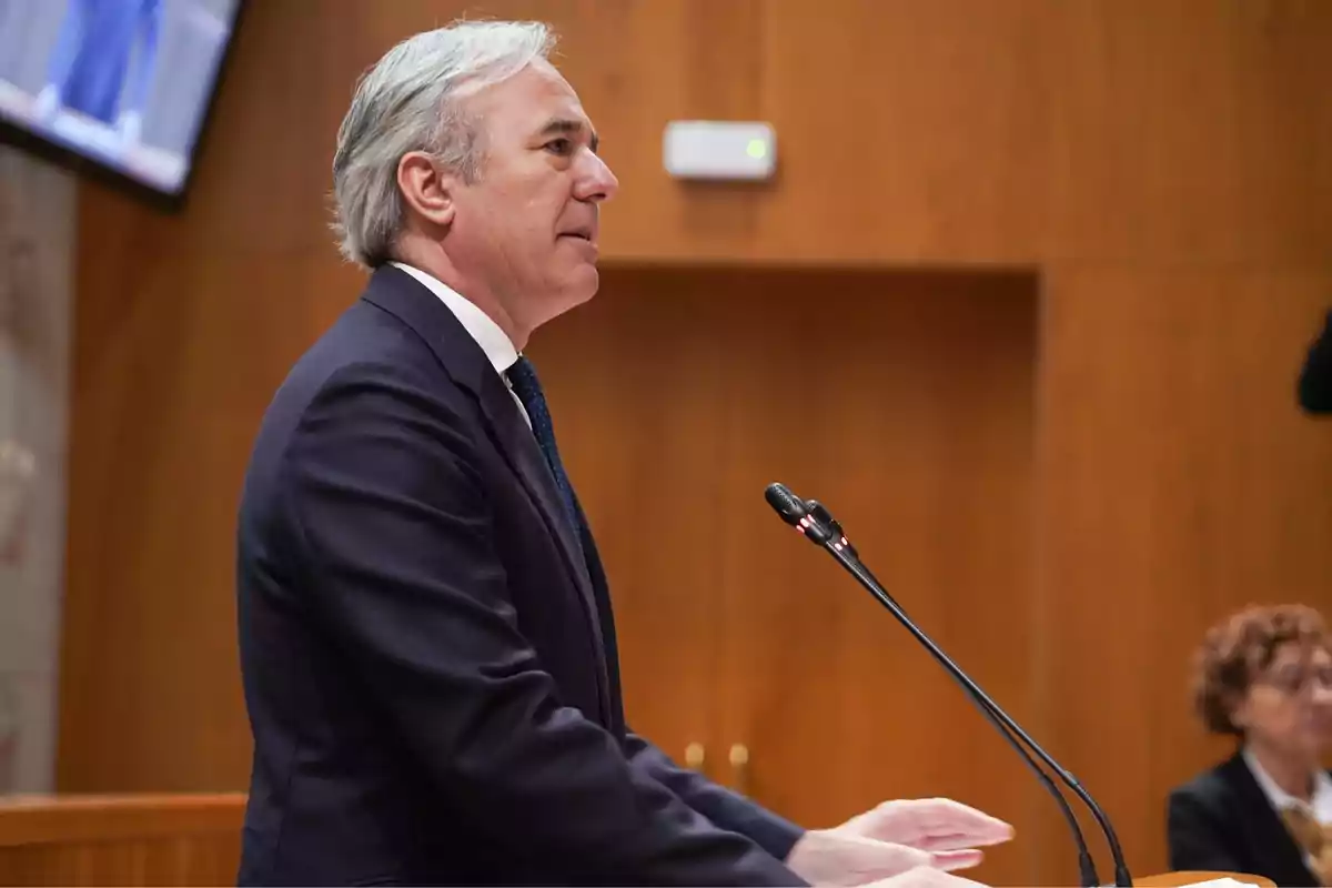 Hombre de cabello canoso hablando en un podio con micrófonos en un entorno formal.