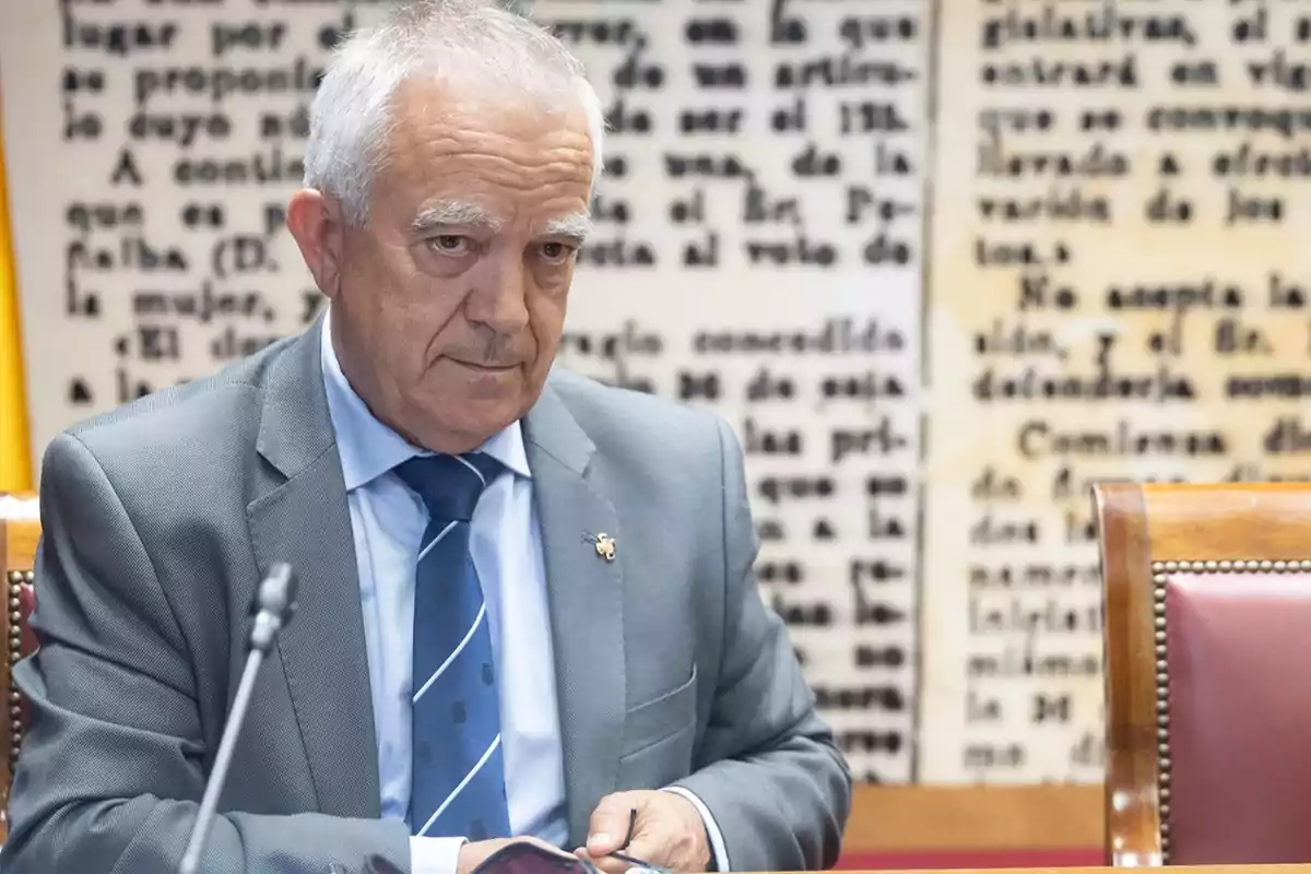 Hombre mayor con traje gris y corbata azul sentado en una sala con fondo de texto borroso.