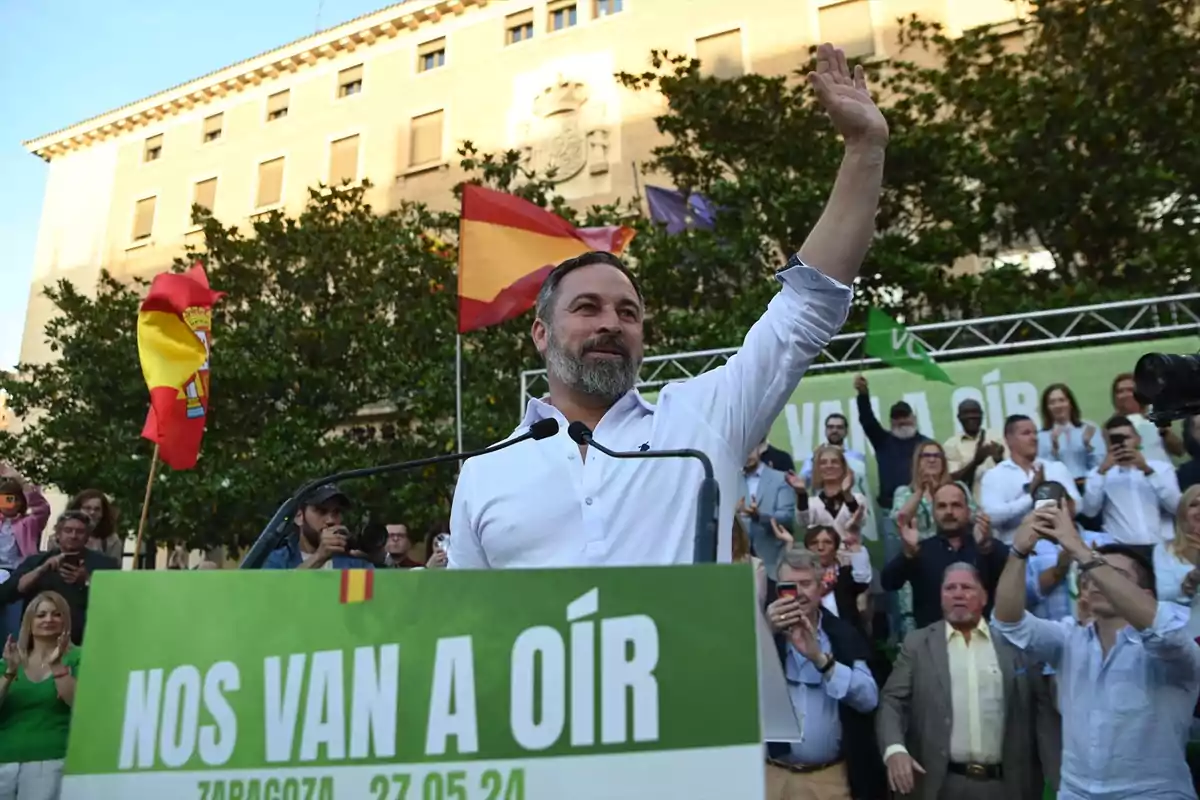 Un hombre con barba y camisa blanca saluda desde un escenario frente a una multitud con banderas españolas y un cartel verde que dice "Nos van a oír" en un evento al aire libre.