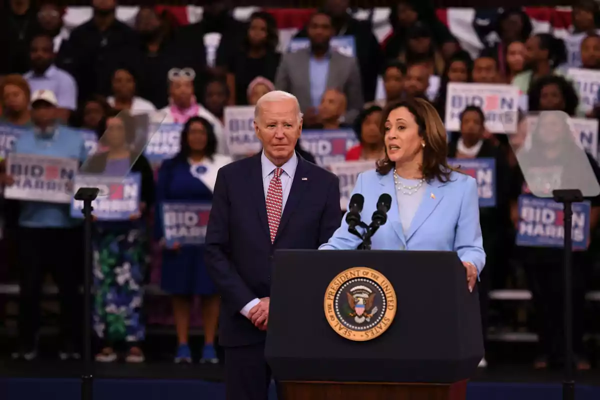 Dos personas hablando en un podio con el sello del Presidente de los Estados Unidos, rodeadas de una multitud que sostiene carteles de apoyo.