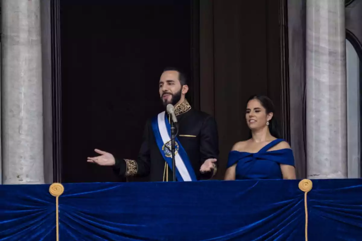 Una pareja vestida de azul oscuro se encuentra en un balcón decorado con tela azul, el hombre está hablando frente a un micrófono.