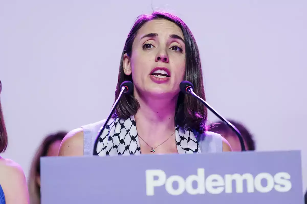 Mujer hablando en un podio con un micrófono y un cartel que dice "Podemos".