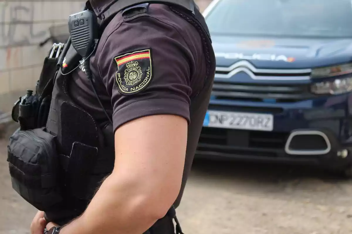 Un agente de la Policía Nacional con uniforme y equipo de comunicación, de pie frente a un vehículo policial.