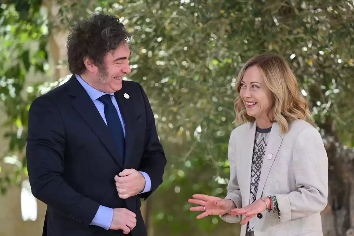 Dos personas elegantemente vestidas conversan y sonríen al aire libre frente a un fondo de vegetación Dos personas elegantemente vestidas conversan y sonríen al aire libre frente a un fondo de vegetación