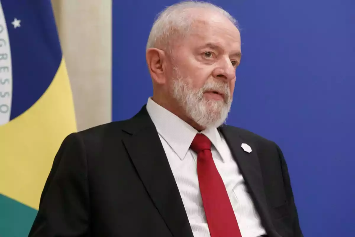 Hombre de barba blanca y traje negro con corbata roja frente a una bandera y un fondo azul.