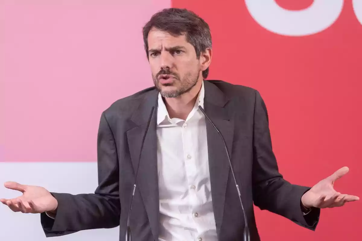 Hombre con traje y camisa blanca gesticulando con las manos mientras habla frente a un micrófono, con un fondo de color rosa y rojo.