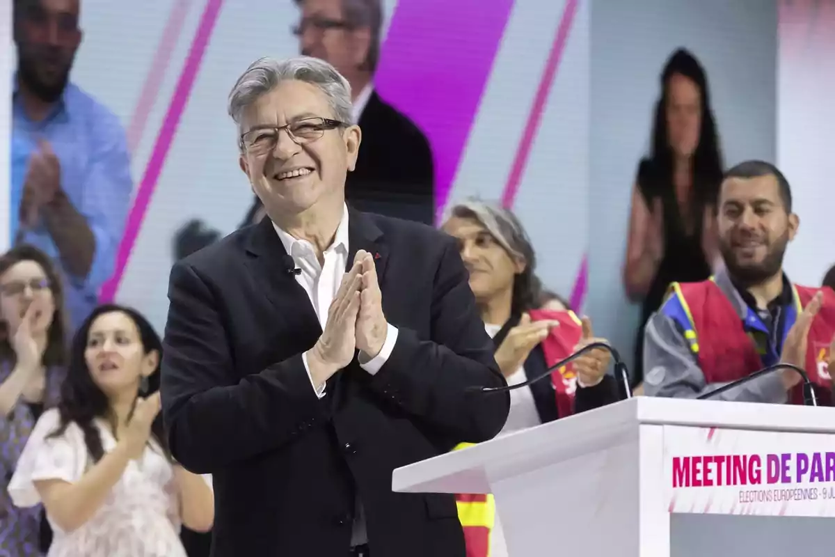 Un hombre mayor con gafas y traje oscuro sonríe y aplaude en un escenario, rodeado de personas que también aplauden, con un cartel que dice "MEETING DE PARIS" en el podio frente a él.