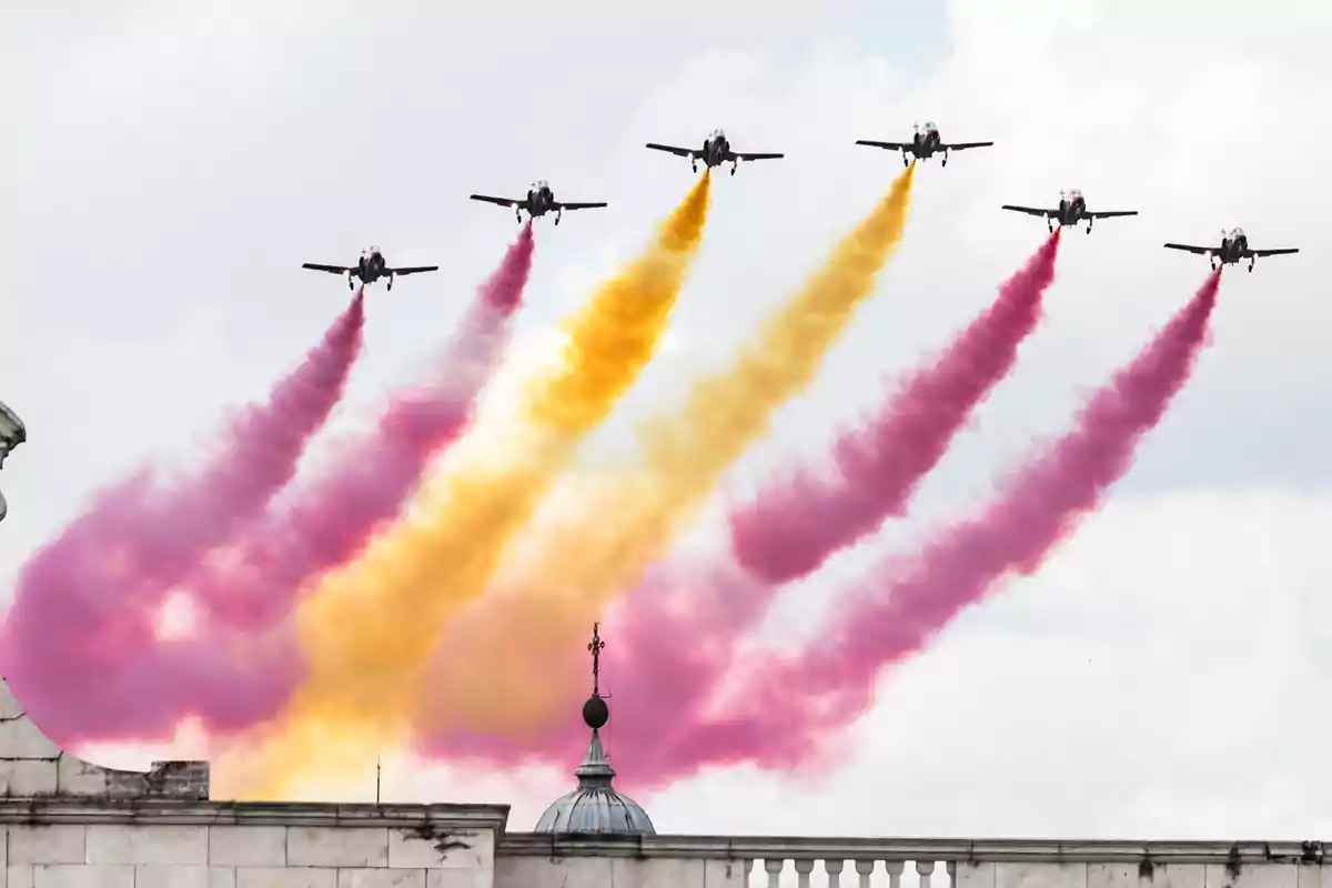 Aviones en formación sobrevolando un edificio mientras emiten humo de colores rojo y amarillo en el cielo.