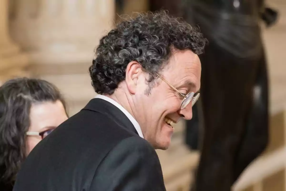 Hombre con gafas y cabello rizado sonriendo mientras viste un traje formal.