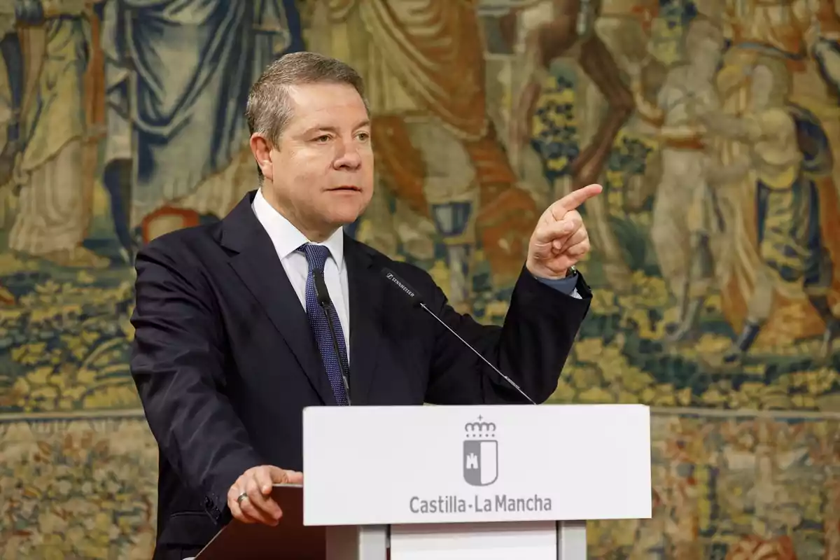 Hombre de traje oscuro y corbata azul hablando en un podio con el escudo de Castilla-La Mancha, señalando con el dedo índice, con un tapiz decorativo de fondo.