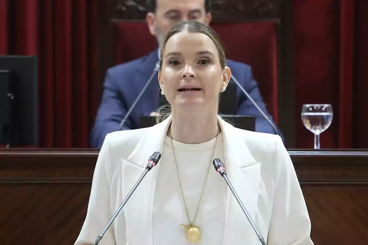 Una mujer hablando en un podio con micrófonos, vestida con un traje blanco y un collar dorado, con una persona desenfocada en el fondo.